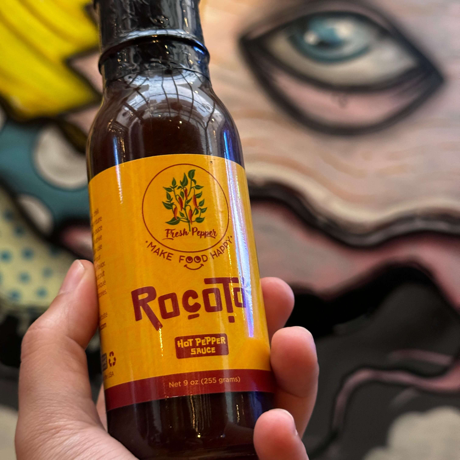 hand holding a bottle of rocoto hot sauce, in the background is a mural